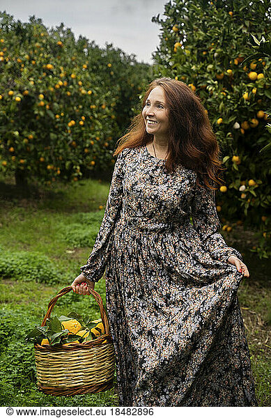 Happy redhead woman standing with basket in front of orange trees