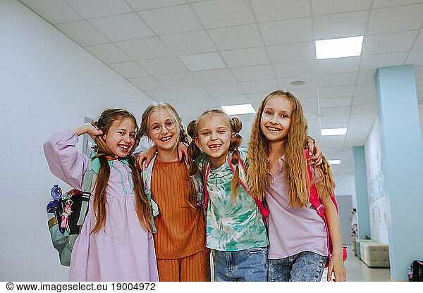 Happy schoolgirls standing with arm around in school corridor