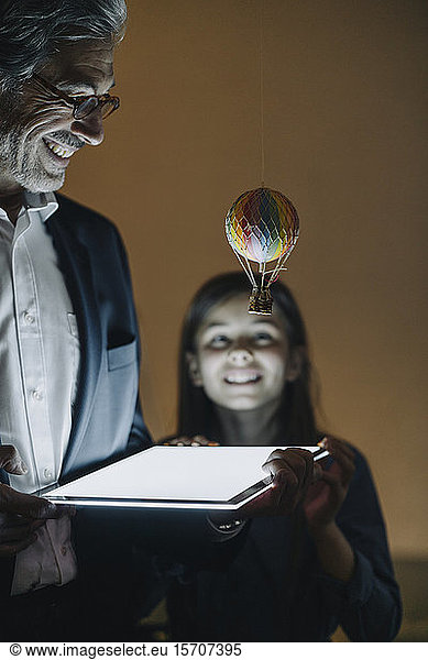 Happy senior buisinessman and girl with hot-air balloon and shining tablet in office