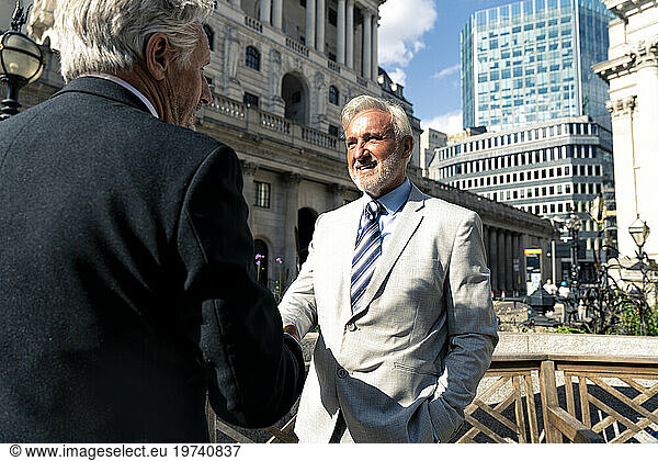 Happy senior businessmen shaking hands together in front of buildings on sunny day