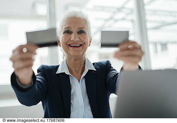 Happy senior businesswoman showing business cards sitting in office