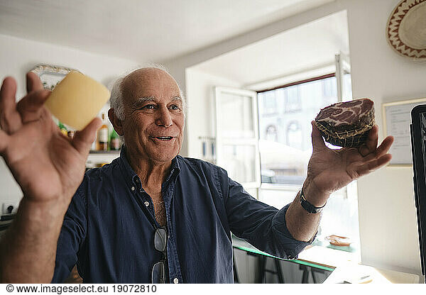 Happy senior cafe owner holding cheese and meat in coffee shop