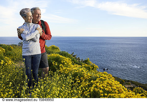 Happy senior couple looking at sea while standing by flowering plants on cliff against sky