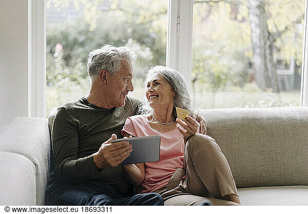 Happy senior couple relaxing on couch at home using tablet for online shopping