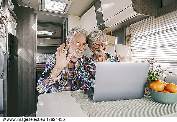 Happy senior couple waving on video call through laptop at motor home