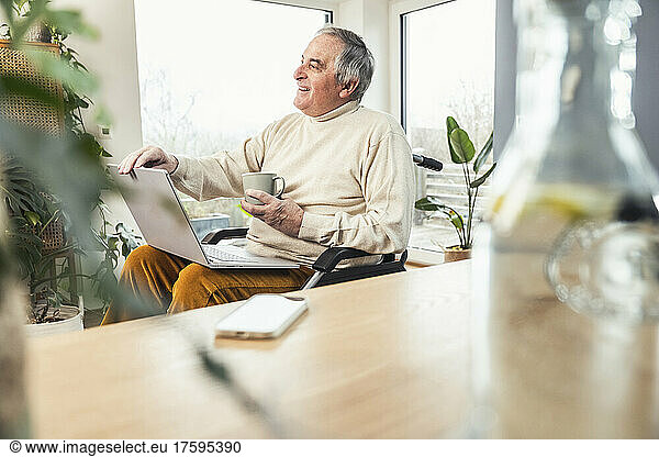 Happy senior disabled man with laptop and coffee cup sitting on wheelchair at home