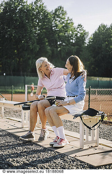 Happy senior female friends talking to each other while sitting on bench at tennis court
