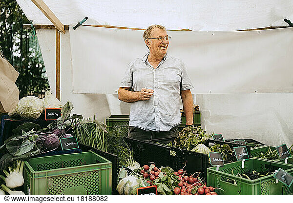 Happy senior male owner looking away while standing amidst vegetables crates at flea market