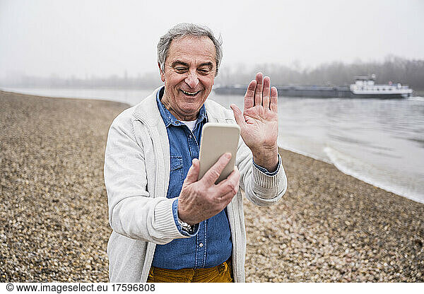 Happy senior man doing video call through smart phone waving hand at beach