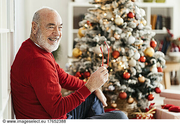 Happy senior man holding eyeglasses sitting in front of Christmas tree at home