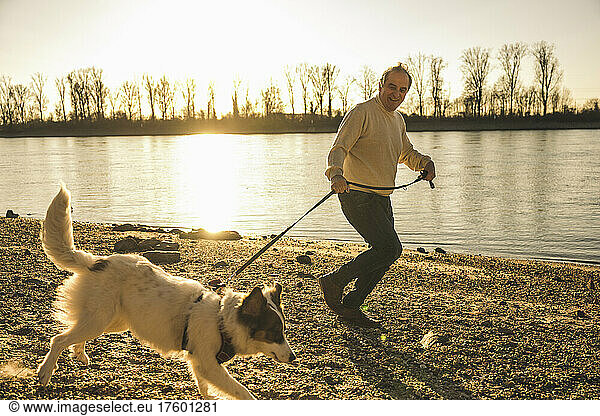 Happy senior man holding pet leash running with dog at beach on sunset