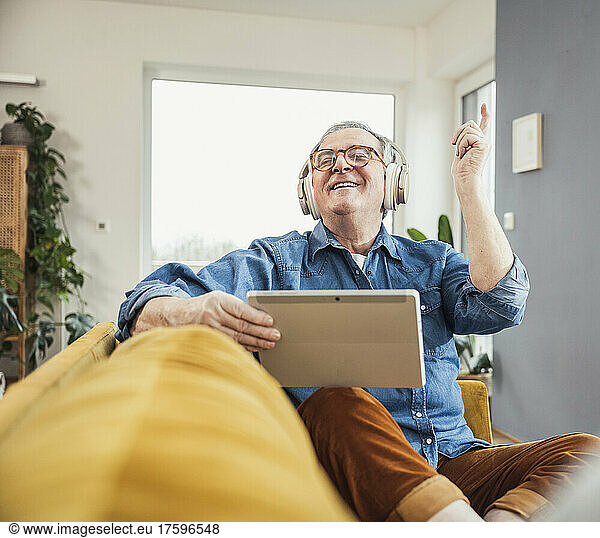 Happy senior man holding tablet PC enjoying music through wireless headphones in living room