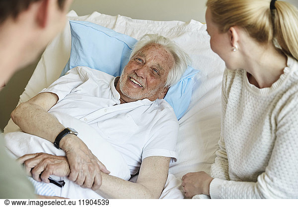 Happy senior man talking to couple while reclining on bed in hospital ward