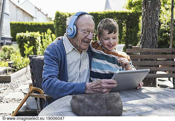 Happy senior man wearing wireless headphones using tablet PC with grandson