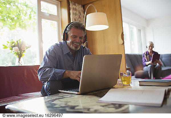 Happy senior man with headphones using laptop in living room