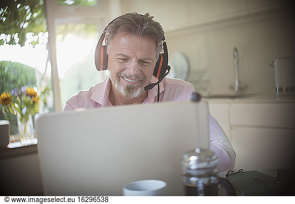 Happy senior man with headphones working at laptop in morning kitchen