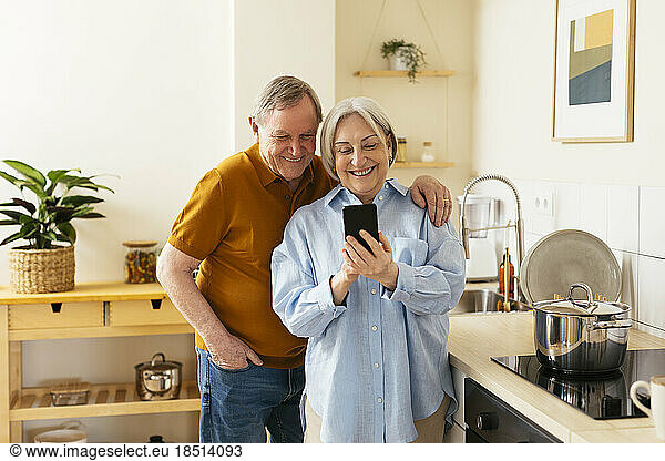 Happy senior woman sharing smart phone with man in kitchen