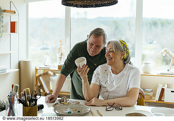 Happy senior woman showing handmade painted cup to man at workshop