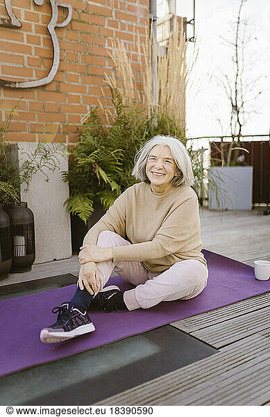 Happy senior woman sitting on exercise mat at terrace