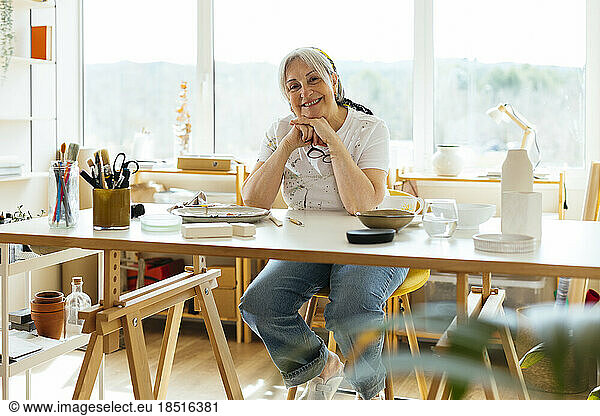 Happy senior woman sitting with hand on chin at table in workshop