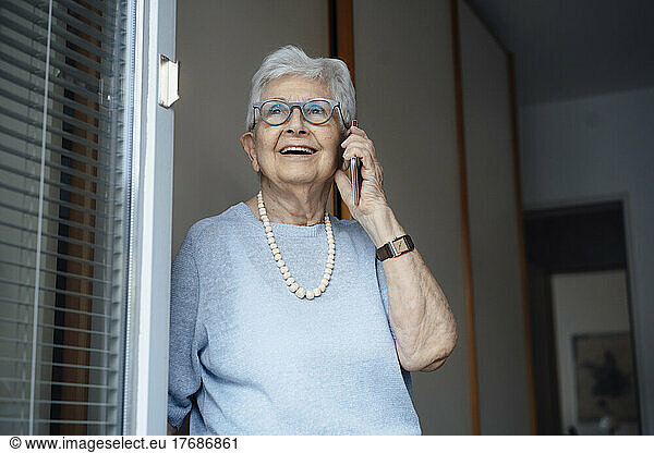 Happy senior woman talking on smart phone standing at apartment doorway