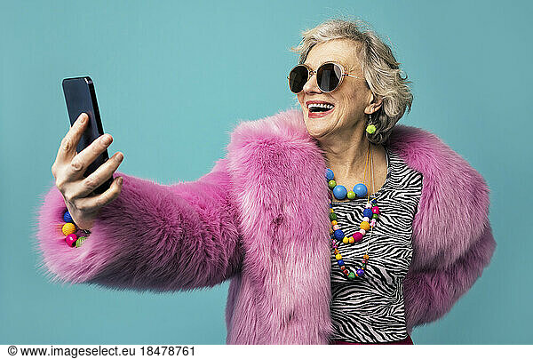 Happy senior woman wearing jewelry taking selfie through smart phone against turquoise background