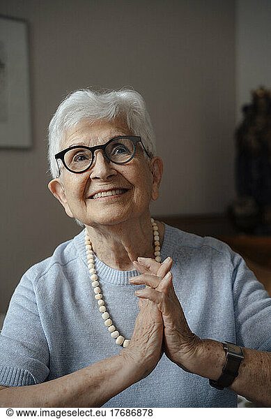 Happy senior woman with hands clasped sitting at home