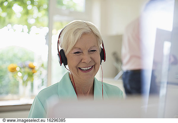 Happy senior woman with headphones using laptop in kitchen