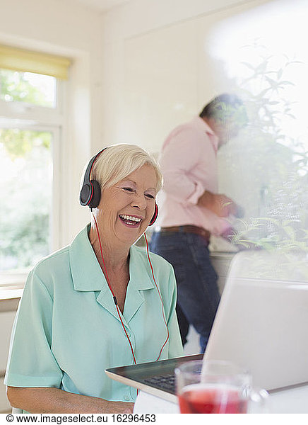 Happy senior woman with headphones video chatting at laptop in kitchen