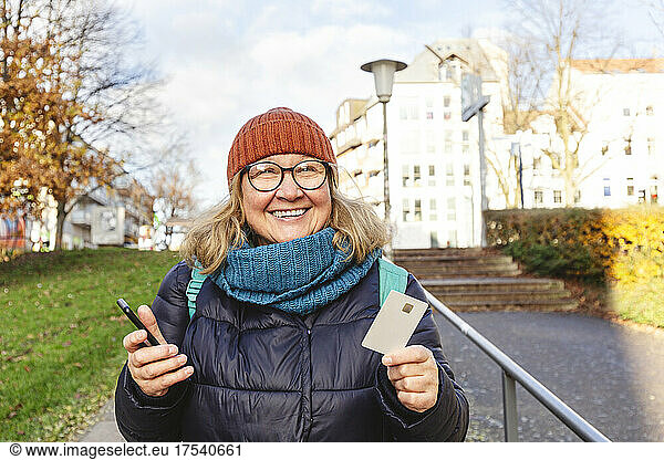 Happy senior woman with smart phone and credit card on footpath