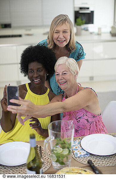 Happy senior women friends in dresses taking selfie at dining table