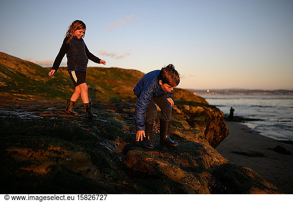 Happy siblings climbing on rocks at sunset near the ocean