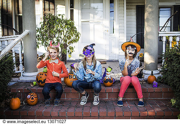 Happy siblings holding props sitting with decorated pumpkins on steps during Halloween