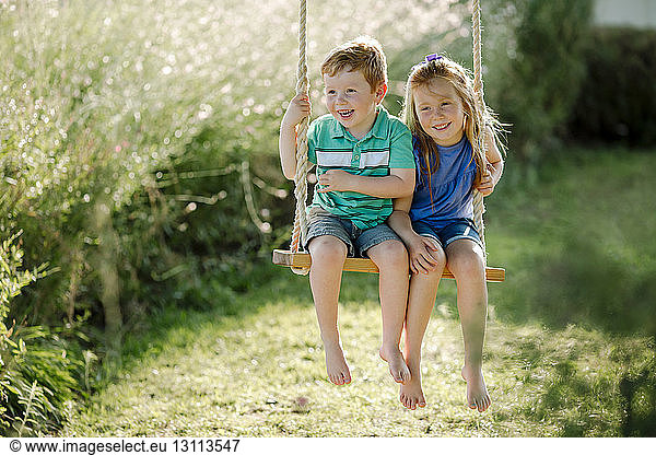 Happy siblings sitting on rope swing at front yard