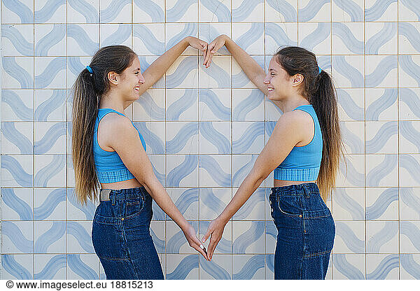 Happy sisters gesturing heart shape in front of wall