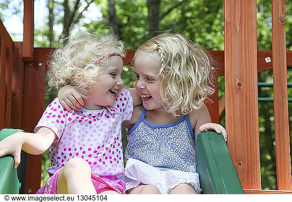 Happy sisters looking face to face while sitting on slide at playground