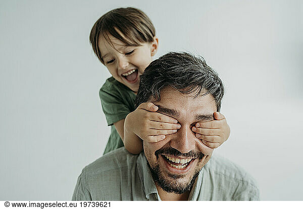 Happy son covering eyes of father against white background