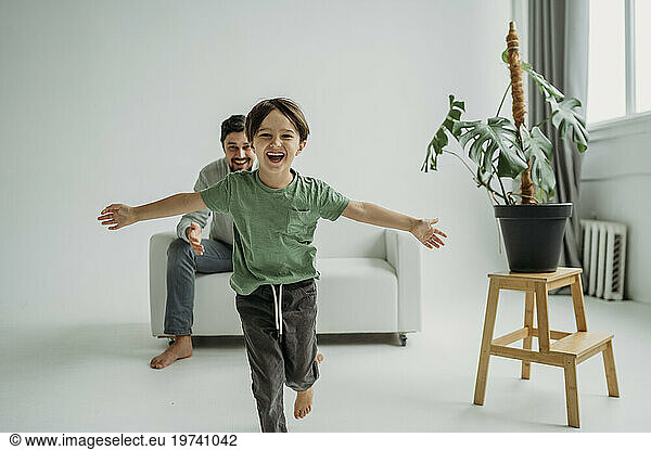 Happy son running with father sitting on sofa in front of white wall