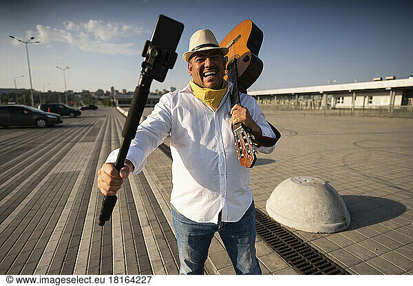Happy street musician holding guitar taking selfie through mobile phone on tripod