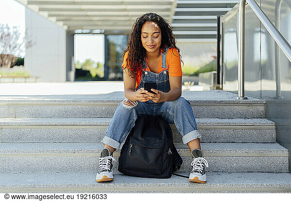 Happy student using smart phone on steps in campus