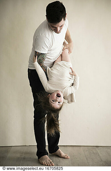 Happy Tattooed Millennial Dad Holds Toddler Daughter Upside Down