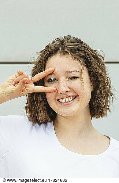 Happy teenage girl winking eye gesturing peace sign in front of wall