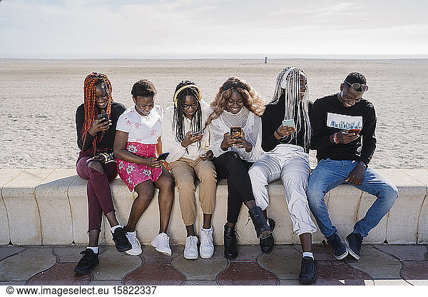 Happy teenage girlfriends and young man sitting on a wall at the beach using smartphones