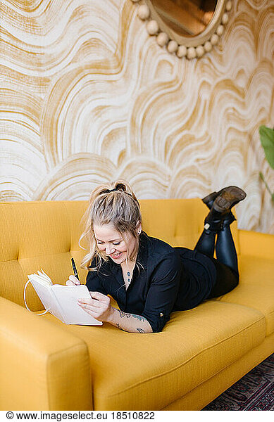 Happy to journal and work  woman laying on bright yellow couch