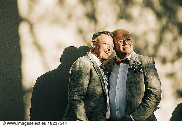 Happy well-dressed gay men standing against wall during wedding on sunny day