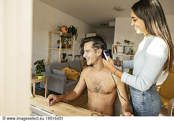 Happy woman cutting boyfriend's hair with electric razor at home