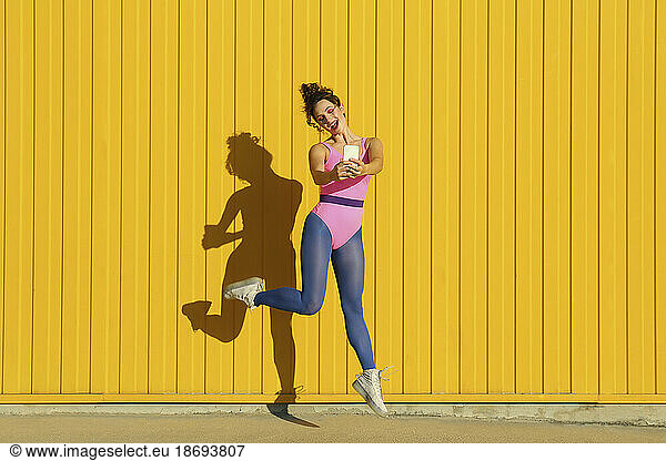 Happy woman dancing and taking selfie through smart phone in front of yellow wall