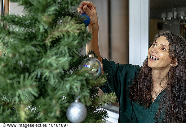 Happy woman decorating Christmas tree with ornaments at home