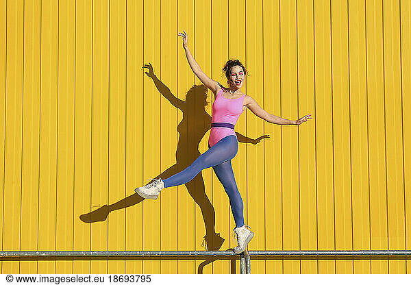 Happy woman doing ballet on railing in front of yellow wall
