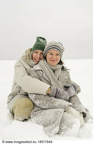 Happy woman embracing friend sitting in snow in winter
